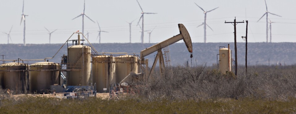 A pumpjack operating on an oil well as wind turbines stand in the